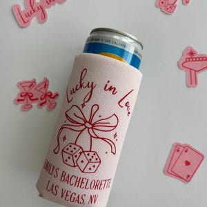 Pode incluir: Resfriador de latas rosa com o texto "Lucky in Love" e uma fita vermelha com dados e cora&ccedil;&otilde;es. O texto "Emily's Bachelorette Las Vegas, NV" est&aacute; impresso abaixo do design.