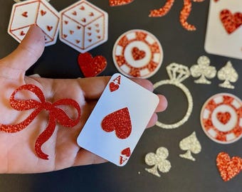 Pode incluir: Vários símbolos de cartas de jogar cobertos com glitter vermelho, dados, corações e fichas de poker. Uma carta de jogar com um coração vermelho e o símbolo do Ás é segurada na mão. Outras formas incluem um laço, um anel e símbolos de cartas.