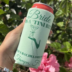 May include: A green and white can cooler with the text "Bride in a "Tini Bit" Jess's Bachelorette Tampa, Fl" printed on it. The cooler is being held by a hand.