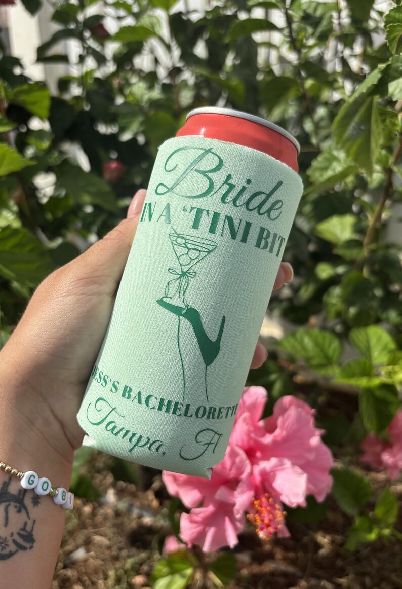 May include: A green and white can cooler with the text "Bride in a "Tini Bit" Jess's Bachelorette Tampa, Fl" printed on it. The cooler is being held by a hand.