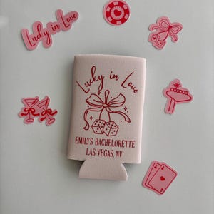 Pode incluir: Frigor&iacute;fico de latas rosa e vermelho com o texto "Lucky in Love" e "Emily's Bachelorette Las Vegas, NV". O refrigerador tem um la&ccedil;o rosa com dados pendurados. H&aacute; v&aacute;rios adesivos rosa e vermelhos ao redor do refrigerador, incluindo um cora&ccedil;&atilde;o, dados, uma ta&ccedil;a de martini, uma ficha de cassino e uma placa de cassino.