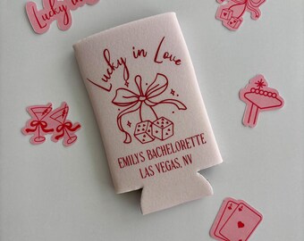 May include: White can cooler with a red design that says "Lucky in Love" and "Emily's Bachelorette Las Vegas, NV". The design also includes a red bow, dice, and a star.