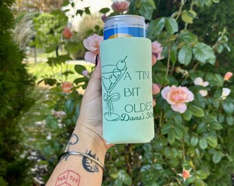 May include: A green and white can cooler with the text "A TINI BIT OLDER Dana's 30th" printed on it. The cooler is being held by a person with a tattoo on their arm.