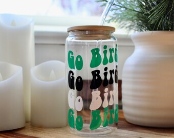 May include: A clear glass water bottle with a bamboo lid and a green, black, and white "Go Birds" design. The bottle is sitting on a wooden surface with candles and a white vase in the background.