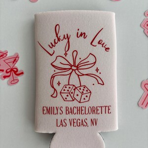 Pode incluir: Resfriador de latas rosa com um design vermelho que diz "Lucky in Love" com um la&ccedil;o e dados. O texto "Emily's Bachelorette Las Vegas, NV" est&aacute; impresso abaixo do design.