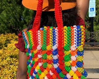 May include: A large, colorful tote bag made of woven plastic beads. The bag is red, orange, yellow, green, blue, and white. The bag has a wide, flat base and a long, rectangular shape. The bag has two long, red handles.