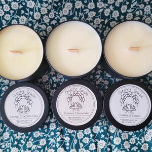 May include: Three black candle jars with white lids and wooden wicks. The labels on the jars read "Gentle Soule" and list the scents "German Chocolate Cake", "Blueberry Cheesecake", and "Cookies & Cream". Each label also states "Handmade in Wisconsin".