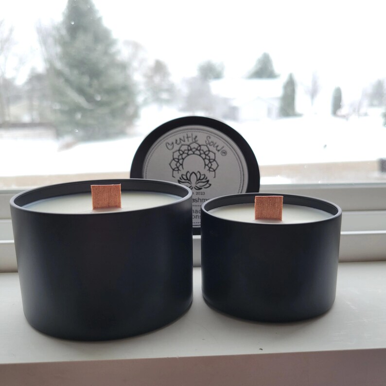 May include: Two black candle jars with wooden wicks. The jars are labeled "Gentle Soul" and have a white label with a mandala design. The candles are sitting on a windowsill.