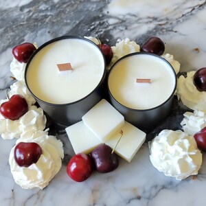 May include: Two black candle jars with white wax and wooden wicks, surrounded by white whipped cream and red cherries.