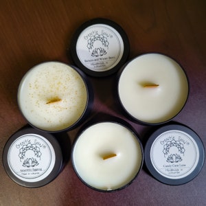 May include: Four black candle jars with white wax and wooden wicks. Each jar has a white label with black text and a floral design. The labels read "Gentle Sowe" and list the scent of each candle: Balsam and Winter Berry, Amaretto Eggnog, Candy Cane Lane.