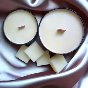 K&ouml;nnte beinhalten: Zwei schwarze Kerzenbeh&auml;lter mit Holzdochten und einer wei&szlig;en Wachsf&uuml;llung. Die Beh&auml;lter stehen auf einem hellvioletten Satin-Stoff mit wei&szlig;en Wachsplatten.