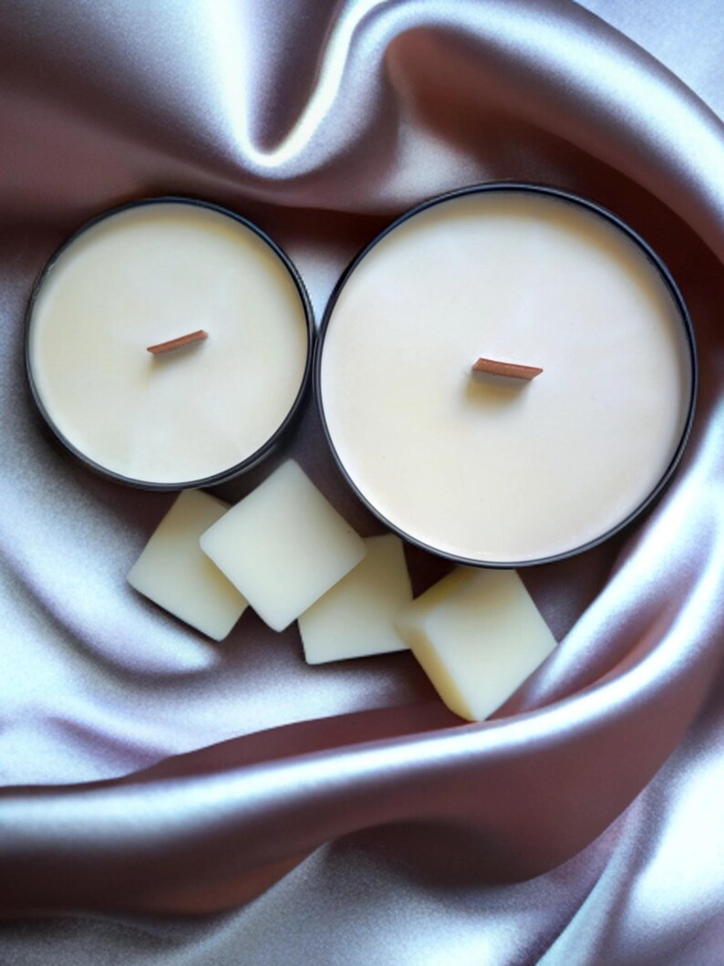 K&ouml;nnte beinhalten: Zwei schwarze Kerzenbeh&auml;lter mit Holzdochten und einer wei&szlig;en Wachsf&uuml;llung. Die Beh&auml;lter stehen auf einem hellvioletten Satin-Stoff mit wei&szlig;en Wachsplatten.