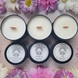 May include: Six black tin candles with white wax and wooden wicks, surrounded by pink, white, and purple flowers. The labels read "Gentle Souls" and list scents like Cedar Rose Cardamom, Midnight Lilac, and Daisy. Handmade in Wisconsin.