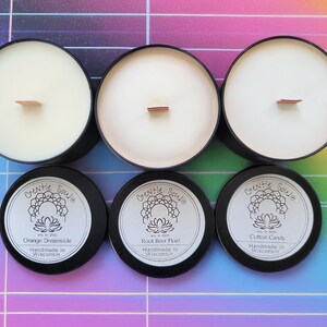 Puede incluir: Tres tarros de velas negros con tapas blancas y mechas de madera. Los tarros están etiquetados con el texto "Gentle Soule" y los aromas "Orange Dreamside", "Root Beer Float" y "Cotton Candy". Los tarros están hechos a mano en Wisconsin.
