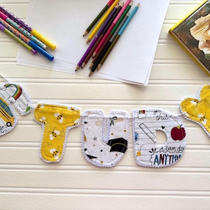 May include: A handmade banner spelling "STUDY" with colorful fabric letters. The letters are decorated with school-themed patterns, including bees, apples, and graduation caps. Pencils, pens, and a book are arranged around the banner on a white surface.
