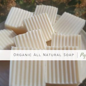 May include: A close-up of a pile of white, organic, all-natural soap bars with a ribbed texture. The soap bars are arranged on a white plate and the text "ORGANIC ALL NATURAL SOAP | Piggy Soap" is visible at the bottom of the image.