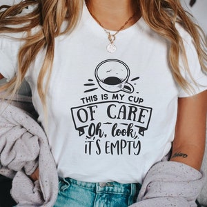 Puede incluir: Camiseta blanca con un diseño gráfico negro que presenta una taza de café y el texto "THIS IS MY CUP OF CARE, Oh, look, IT'S EMPTY." La camiseta es una prenda informal.