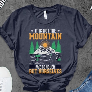 Puede incluir: Una camiseta azul marino con una escena de montaña y el texto "No es la montaña la que conquistamos, sino nosotros mismos".