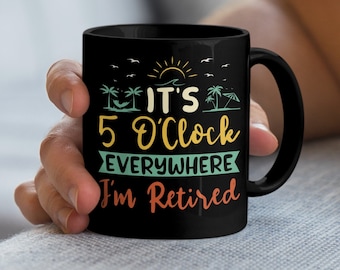 Op de afbeelding: Zwarte koffiemok met een witte afbeelding waarop staat "It's 5 O'Clock Everywhere I'm Retired". De afbeelding bevat ook een zon, palmbomen en een parasol.