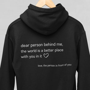 Op de afbeelding: Zwart hoodie met de tekst "dear person behind me, the world is a better place with you in it" en een hartje. De tekst "love, the person in front of you" staat onder het hartje.