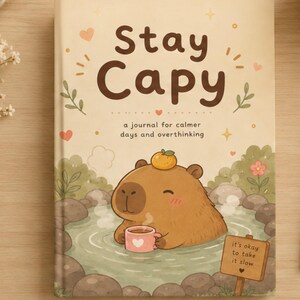May include: A hardcover journal with a cream-colored cover featuring a brown capybara in a hot spring with an orange on its head, holding a pink mug. The text reads Stay Capy, a journal for calmer days and overthinking, and it's okay to take it slow.