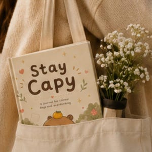 May include: A cream-colored hardcover journal with the text Stay Capy and a small illustration of a capybara with an orange on its head. The journal is tucked inside a beige canvas tote bag alongside a small bouquet of white baby's breath flowers.