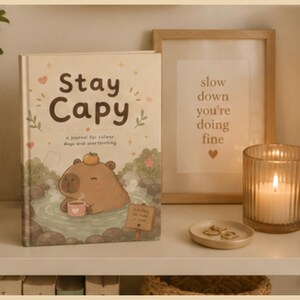 May include: A hardcover journal titled Stay Capy with a cream-colored cover featuring an illustration of a capybara in a hot spring, holding a mug with a small orange on its head, surrounded by small hearts, stars, and green leaf accents.