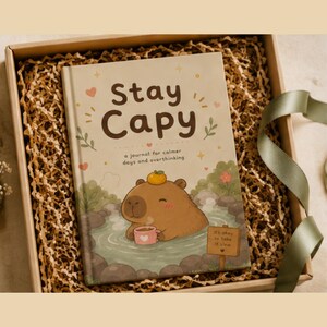 May include: A hardcover journal titled Stay Capy: a journal for calmer days and overthinking. The cover features a brown capybara in a hot spring with a small orange on its head, holding a pink mug, with a sign reading it's okay to take it slow.