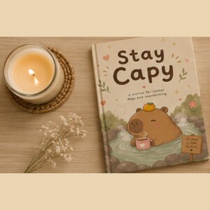 May include: A hardcover journal titled Stay Capy with a brown capybara illustration in a hot spring, holding a pink mug with a heart. The cover features text reading Stay Capy, a journal for calmer days and overthinking, and It's okay to take it slow.