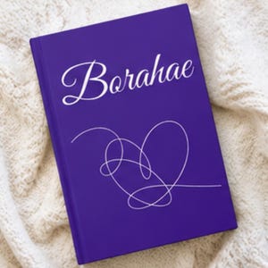 Può includere: Un quaderno viola con la parola "Borahae" in corsivo bianco e un disegno a forma di cuore bianco. Il quaderno è appoggiato su una superficie bianca e strutturata.