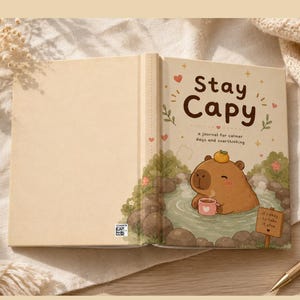 May include: A beige journal titled Stay Capy, a journal for calmer days and overthinking. The cover features a brown capybara with an orange on its head, sitting in a hot spring holding a pink mug. A sign reads it's okay to take it slow.