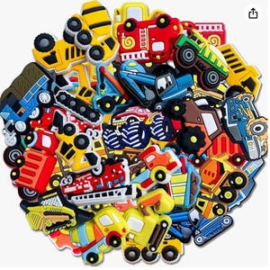 Könnte beinhalten: Eine Sammlung bunter Schuhanhänger aus Gummi in Form verschiedener Baufahrzeuge, Feuerwehrautos, Monstertrucks und Bagger. Das Sortiment umfasst leuchtend gelbe, rote, blaue und grüne Designs mit detaillierten schwarzen Rädern und mechanischen Teilen.