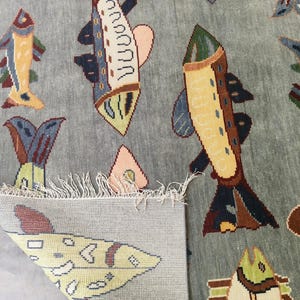 May include: A light blue rug featuring a repeating pattern of colourful fish. The fish are in shades of yellow, blue, brown, and pink. The rug has a fringe along the edge.