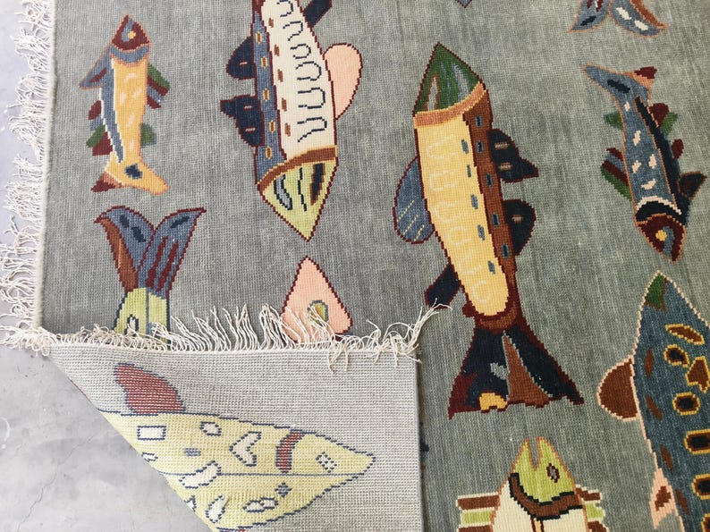 May include: A light blue rug featuring a repeating pattern of colourful fish. The fish are in shades of yellow, blue, brown, and pink. The rug has a fringe along the edge.