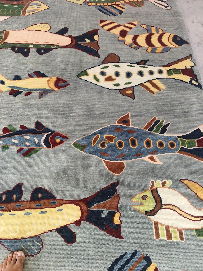 May include: A light blue rug with a repeating pattern of colourful fish. The fish designs feature various colours, including yellow, brown, blue, and green, creating a vibrant and playful aesthetic. The rug is likely made of woven material.