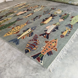 May include: A rectangular, pale blue rug featuring a repeating pattern of colourful fish. The fish vary in size and design, with shades of yellow, green, blue, and red. The rug has a fringed edge.