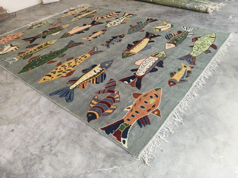 May include: A rectangular, pale blue rug featuring a repeating pattern of colourful fish. The fish vary in size and design, with shades of yellow, green, blue, and red. The rug has a fringed edge.