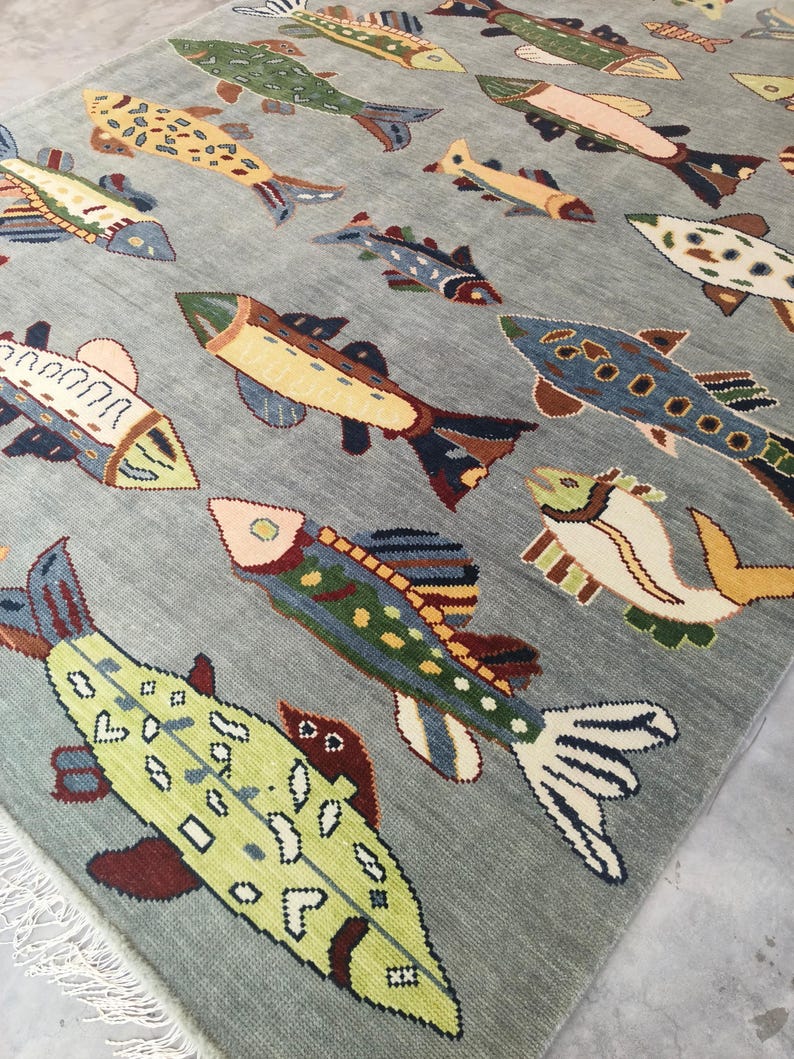 May include: A rectangular rug with a light grey background displaying a repeating pattern of colourful fish. The fish vary in size and colour, including shades of green, yellow, blue, and brown. The rug has a fringe along one edge.