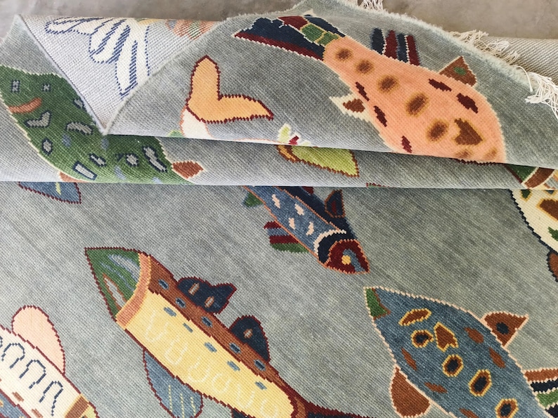 May include: A pale blue rug featuring a repeating pattern of fish in various colours. The fish are orange, blue, green, and yellow, with brown and white details. The rug has a textured finish.