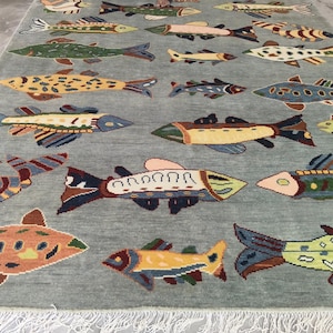 May include: A rectangular rug featuring a light blue background adorned with a repeating pattern of vibrant fish. The fish are depicted in shades of orange, yellow, green, and blue, with detailed fins and tails. The rug has a fringe along the bottom edge.
