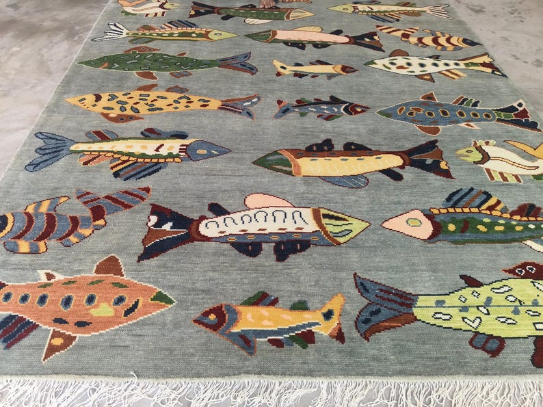 May include: A rectangular rug featuring a light blue background adorned with a repeating pattern of vibrant fish. The fish are depicted in shades of orange, yellow, green, and blue, with detailed fins and tails. The rug has a fringe along the bottom edge.