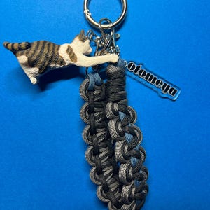 以下が含まれることがあります： 茶色と白の模様の猫の置物と、黒、グレー、青色の編み込みストラップが付いたキーホルダー。キーホルダーにはシルバーのリングと「otomeya」の文字が入った透明なタグが付いています。