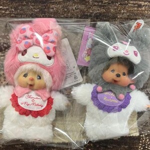 以下が含まれることがあります： プラスチック包装に入った、ピンクとグレーの2つのぬいぐるみハンドパペット。ピンクのパペットはイチゴ模様のリボンと「Chintan My Melody」と書かれたよだれかけが付いています。グレーのパペットにはリボンと「Monchhichi Kuromi」と書かれたよだれかけが付いています。