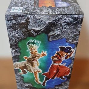 May include: A box featuring the Dr. Stone anime series. The box art shows two characters in action poses against a green and blue background. The text "Dr. Stone FIGURE of STONE WORLD" is visible, along with Japanese text and the date "2023年4月登場予定!"