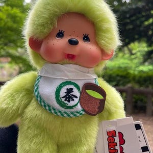 May include: A plush green Monchhichi doll with a matching head covering and bib. The bib features a green circle with a Japanese tea symbol and a brown teacup with green tea. The doll has rosy cheeks and a black nose.