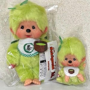 Op de afbeelding: Twee groene pluche poppen, een grote en een kleine, in een transparante plastic verpakking. De poppen hebben groen haar, sproeten en slabbetjes met een theekopjesontwerp.