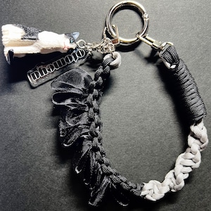 Può includere: Un portachiavi in paracord nero e grigio con chiusura argentata e una piccola statuina di gatto. Il portachiavi presenta una sezione in nastro nero arricciato e un ciondolo argentato con la scritta "otomoya".