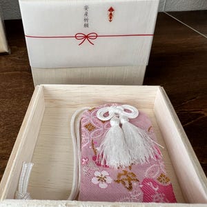 以下が含まれることがあります： 淡い木箱に入ったピンク色の布製お守り。白い桜の模様と金色の刺繍が施されています。箱の蓋は白い紙で包まれ、赤い紐と「安産祈願」という日本語の文字が記されています。