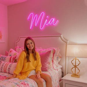 Puede incluir: Un letrero de ne&oacute;n rosa brillante montado en una pared que muestra el nombre Mia en una fuente cursiva. El letrero emite un brillo vibrante, iluminando el espacio sobre una cama con cabecera blanca, ropa de cama floral y una l&aacute;mpara de mesa dorada.