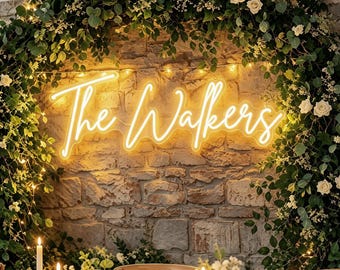 May include: A bright yellow neon sign displaying the text The Walkers in a cursive script font, mounted on a rustic stone wall framed by a large circular arch of lush green foliage, white roses, and delicate string lights above a table.
